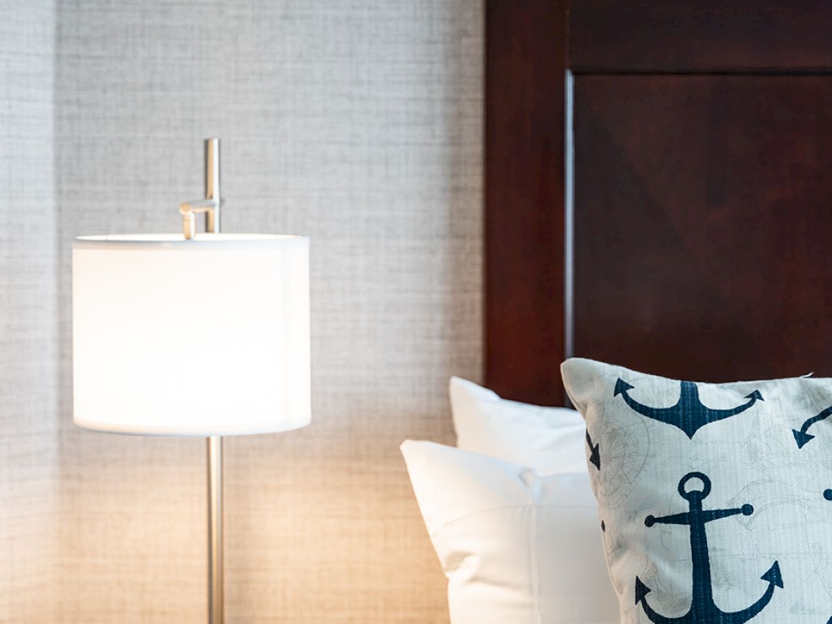 A bedside setup with a wooden headboard, a white pillow, an anchor-themed pillow, a nightstand, and a modern table lamp with a white shade ends the sentence.