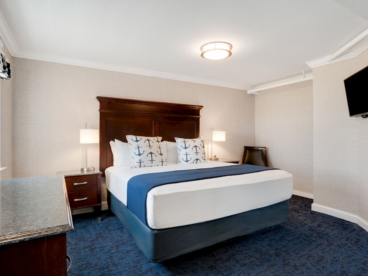 A well-lit hotel room featuring a double bed with nautical-themed pillows, nightstands with lamps, a wall-mounted TV, and a window with a view.