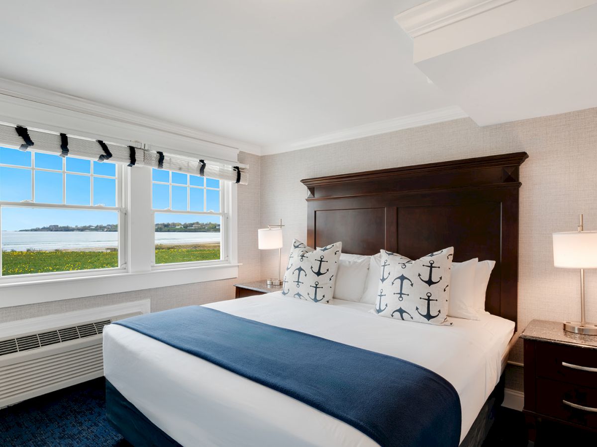 A bedroom with a large bed, nautical-themed pillows, and a blue blanket, featuring two bedside tables with lamps, and a window with a scenic view.