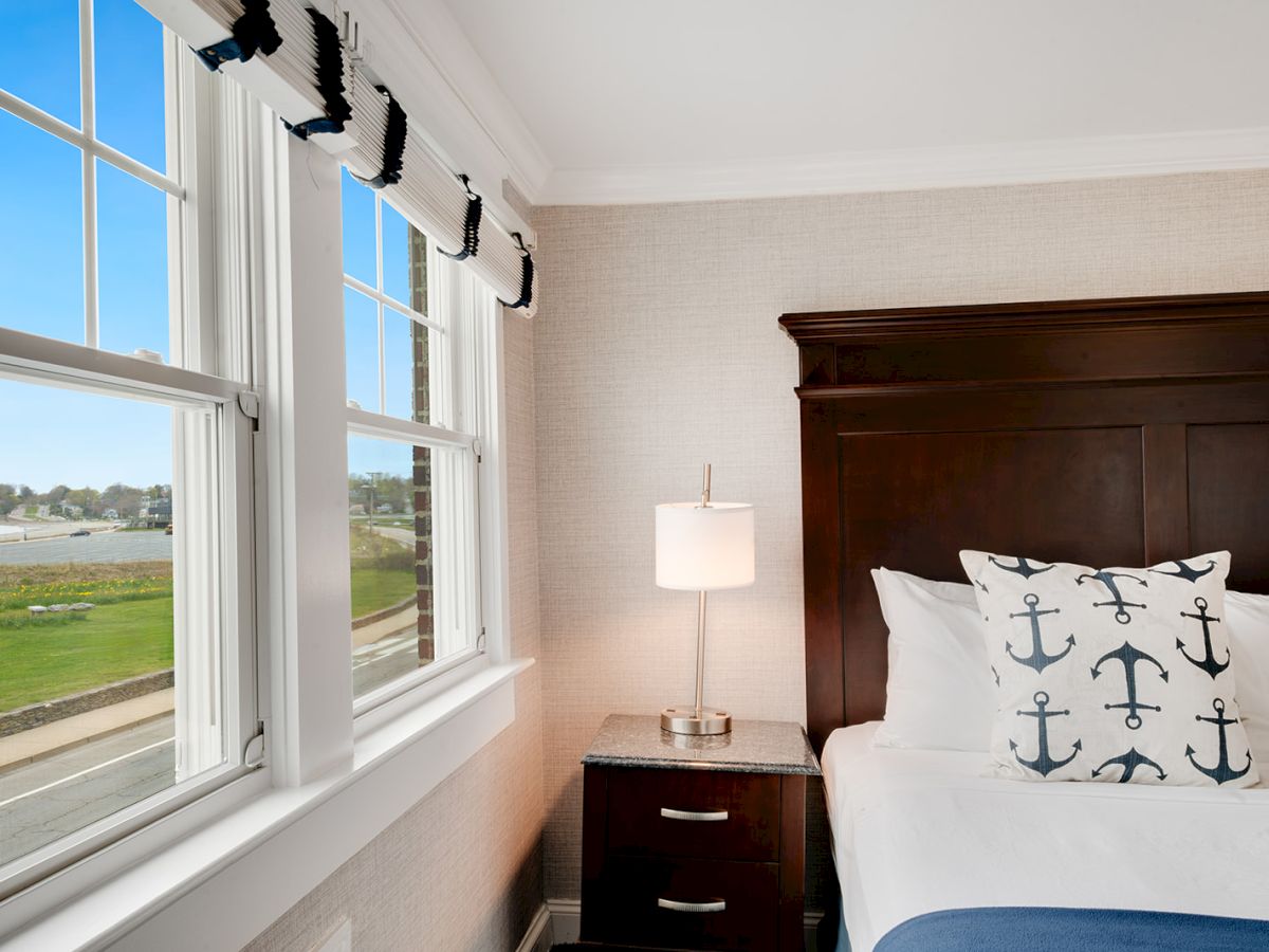 A cozy bedroom with nautical-themed pillows, beside a bedside lamp and table, and large windows showcasing a scenic outdoor view.