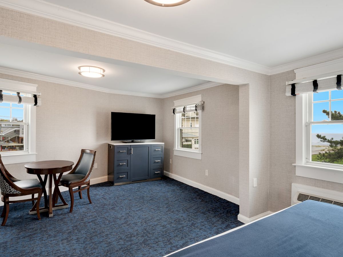 An image of a room with a small table and chairs, a TV on a dresser, and multiple windows with scenic views, having light-colored walls and blue carpet.