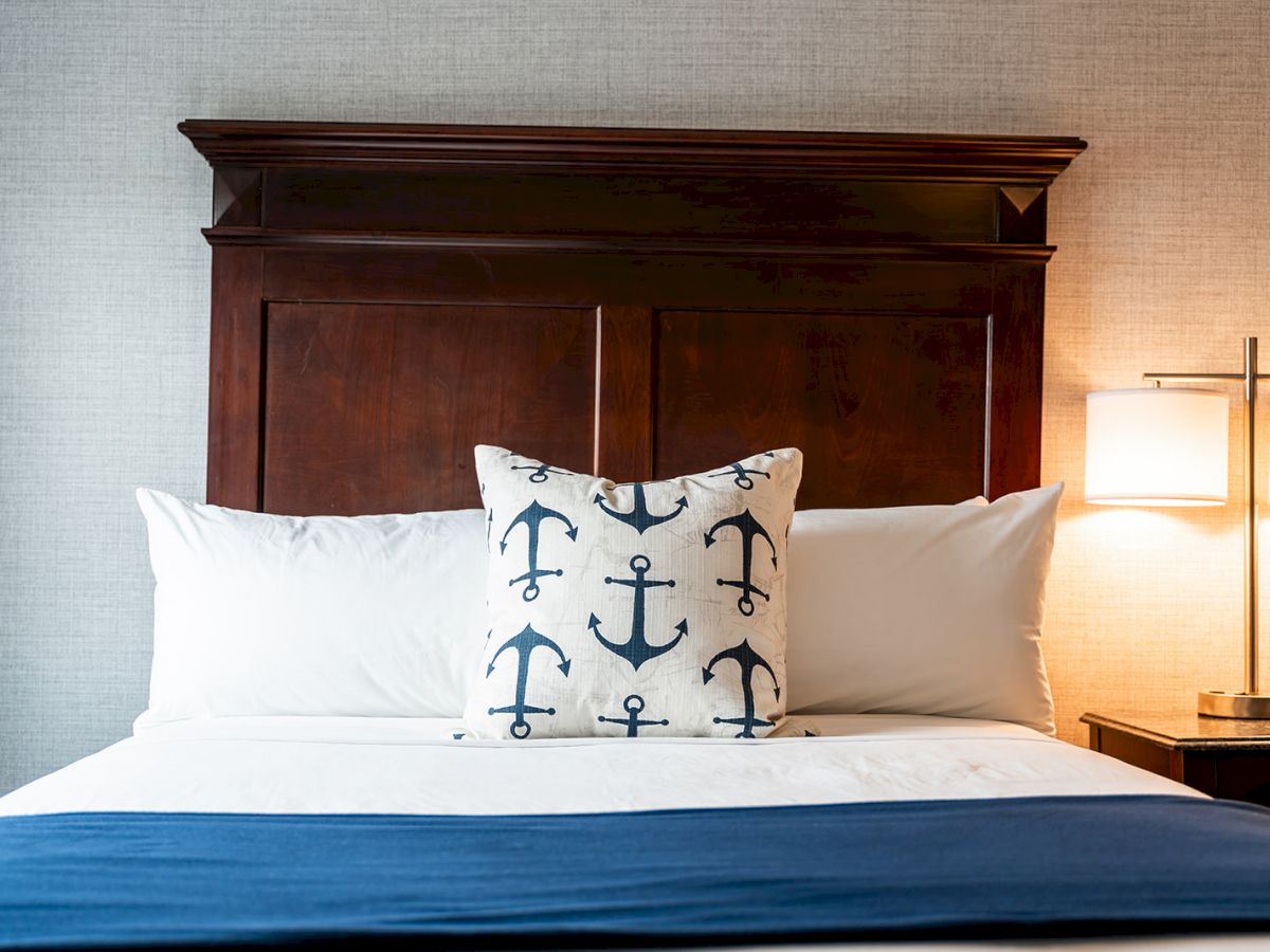 The image shows a neatly made bed with white linens, a blue blanket, and a decorative pillow with anchor patterns, next to a lit bedside lamp.