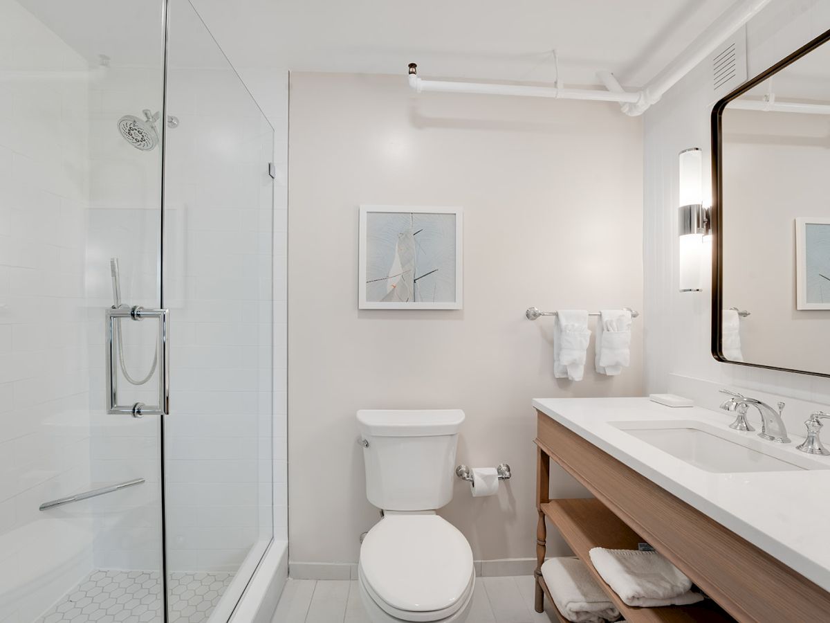 This image shows a modern bathroom with a glass shower, toilet, large mirror, and countertop with sink, along with towels and framed artwork on the wall.