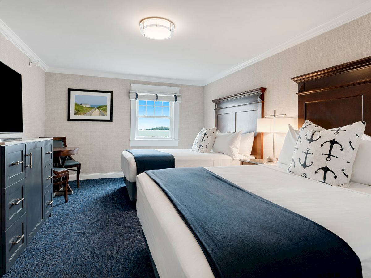 A hotel room with two neatly made beds featuring anchor-themed pillows, a large TV, a small table with chairs, a window, and framed artwork.