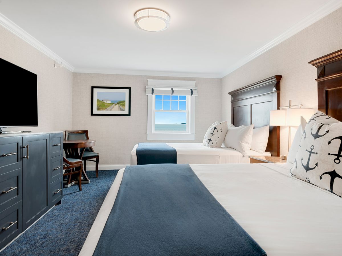 A hotel room with two beds, nautical-themed pillows, a TV, a dresser, a desk, and a window with a scenic view.