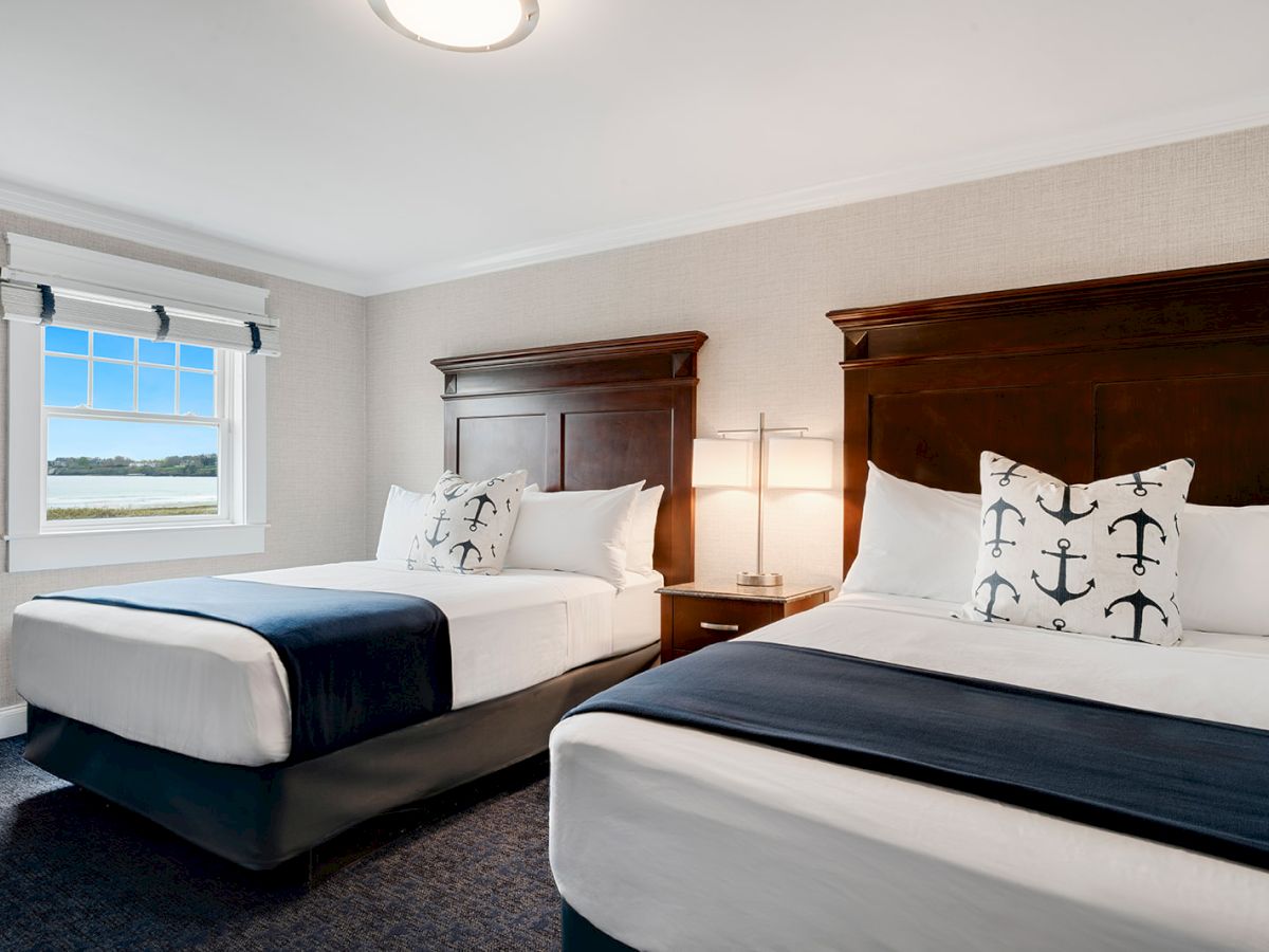 A bright, clean hotel room features two neatly made beds with white linens and dark wood headboards, plus nautical-themed pillows and a scenic window view.