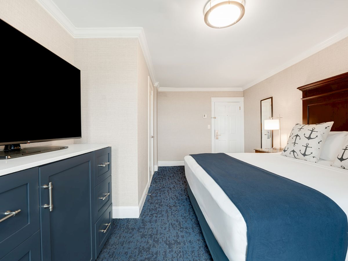The image shows a hotel room with a bed, nautical-themed pillows, a blue and white color scheme, a TV on a dresser, and a door in the background.