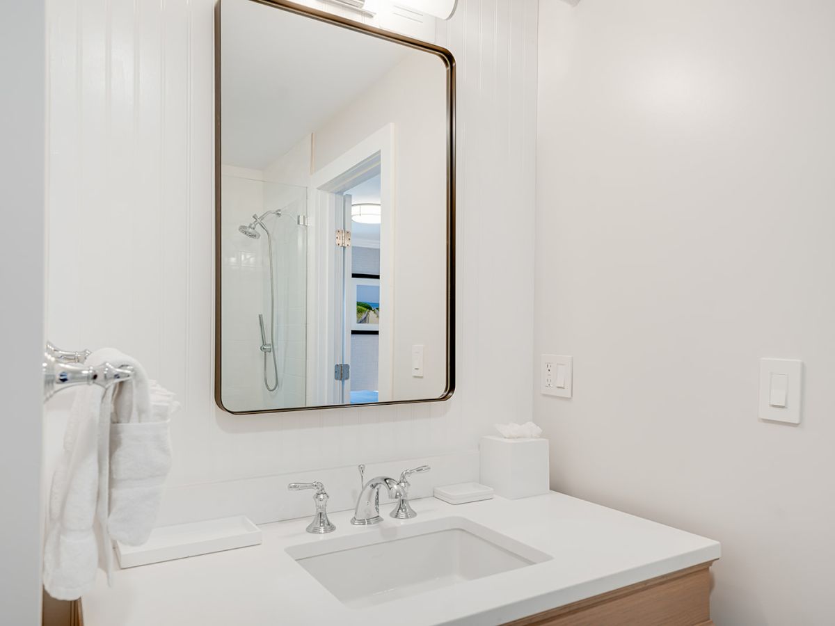 A bathroom with a sink, mirror, and overhead light. Towels on a rack, a tissue box, and a shower is seen in the reflection.
