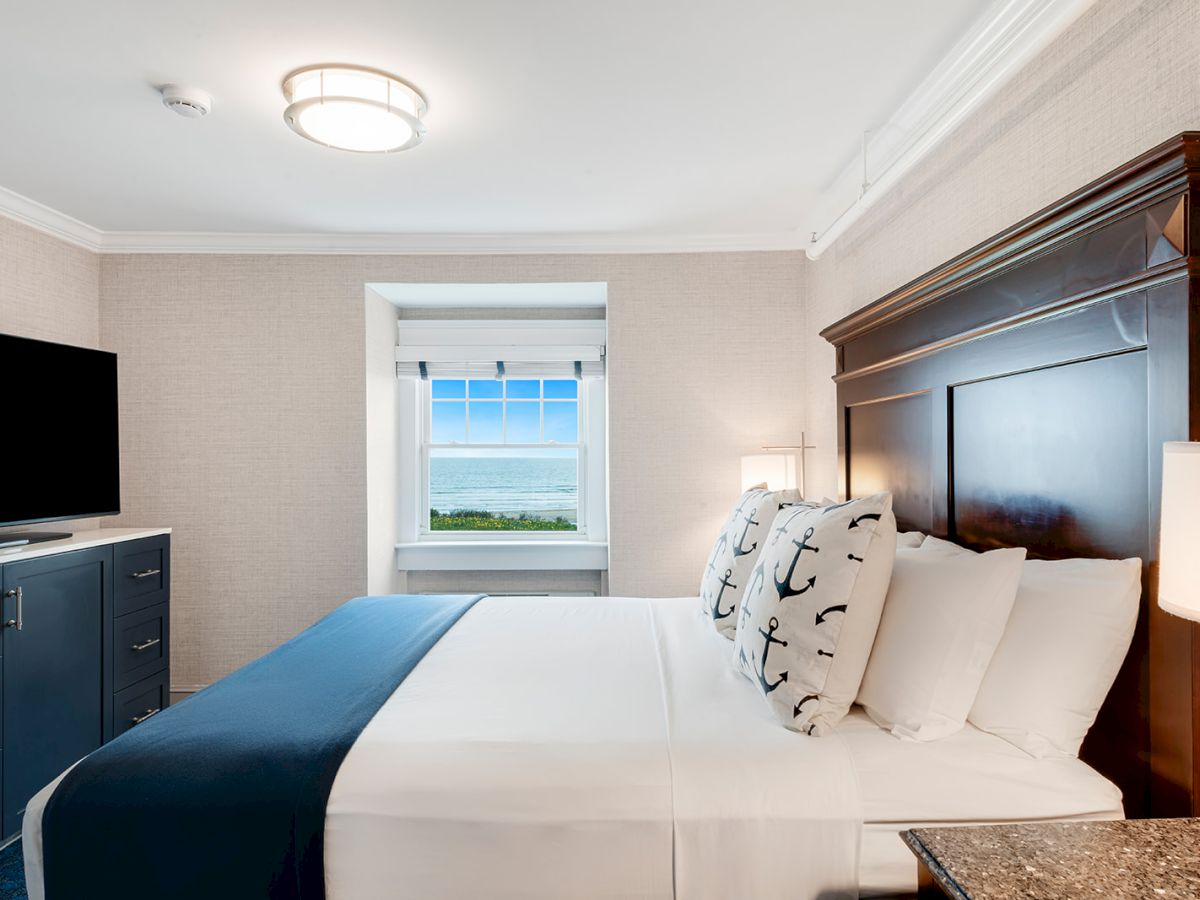 A tidy bedroom with a bed, pillows, dark wooden headboard, TV on a cabinet, a window with an ocean view in the background, and a light overhead.