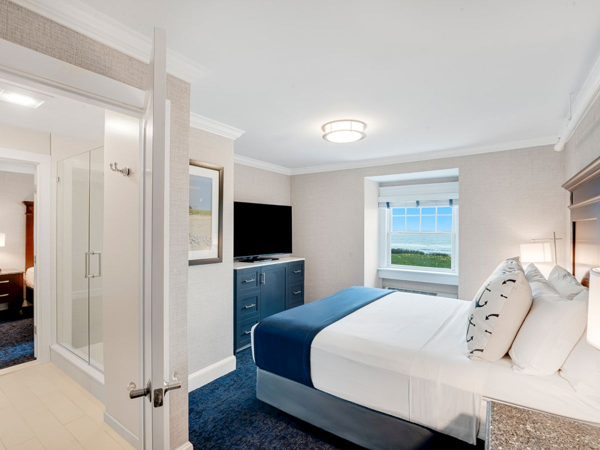 This image shows a modern bedroom with a neatly made bed, a flat-screen TV, and a view of the coast through the window. The door leads to a bathroom.