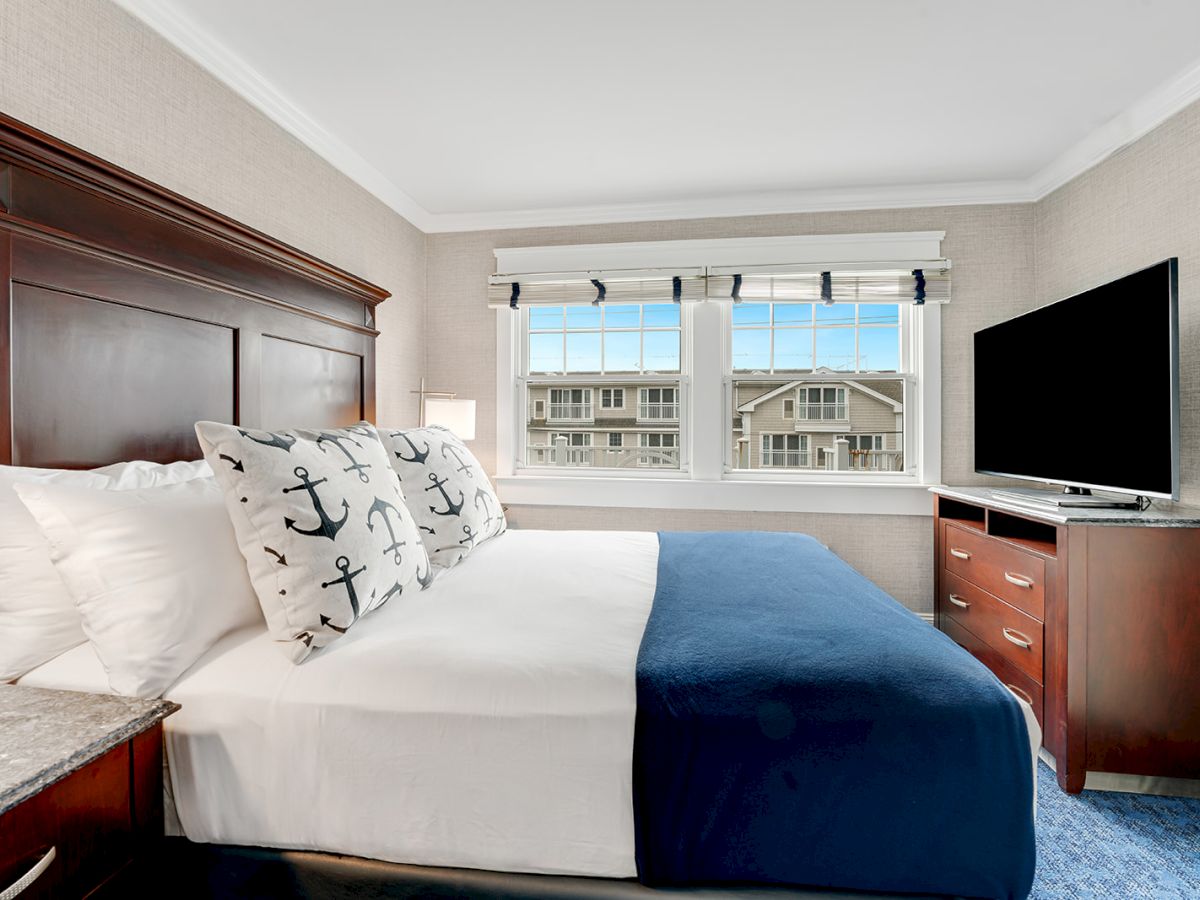 A modern, neatly arranged bedroom features a bed with anchor-themed pillows, a blue blanket, a large flat-screen TV, and a double window.