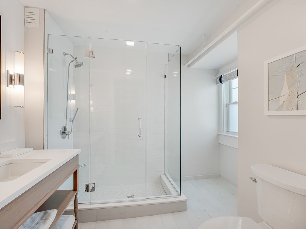 The image shows a modern, white bathroom with a glass shower, a sink with a mirror, a toilet, and a wall-mounted light fixture.