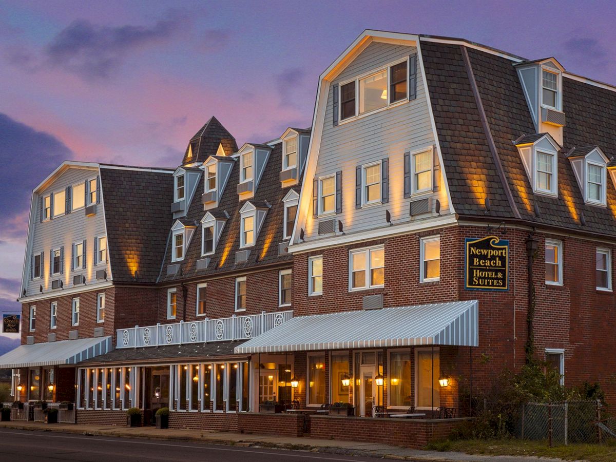 A multi-story brick building with a sign reading "Newport Beach Hotel & Suites" is shown at dusk with lights on inside and outside.