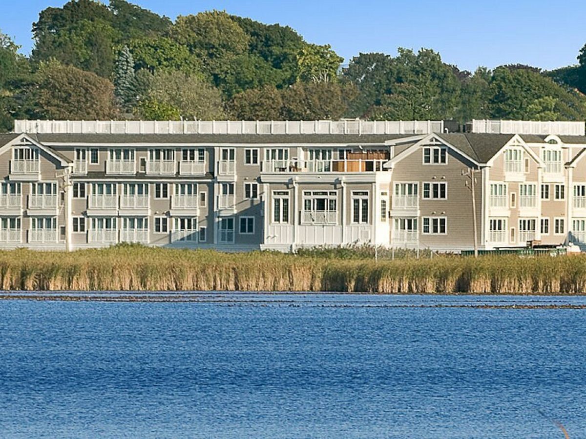 The image shows a large, light-colored building near a body of water, with lush green trees in the background, under a clear blue sky.