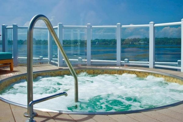 A circular hot tub with bubbling water, set on a deck with clear glass railings overlooking a scenic body of water under a blue sky.