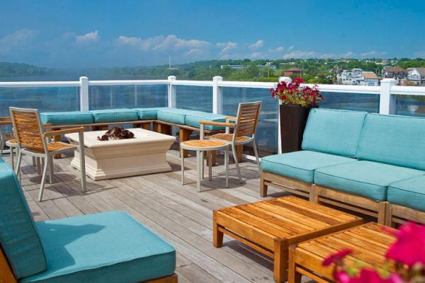 A rooftop patio with blue cushioned seating, wooden tables, a fire pit, and flowers, overlooking a body of water with buildings in the background.