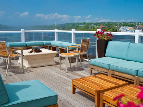 A rooftop patio with blue cushioned seating, wooden tables, a fire pit, and flowers, overlooking a body of water with buildings in the background.