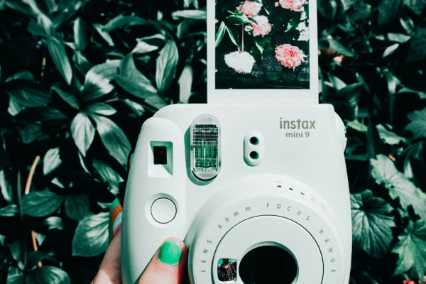 A hand with green nail polish is holding a white Instax camera, displaying a photo of flowers, against a background of lush green leaves and flowers.