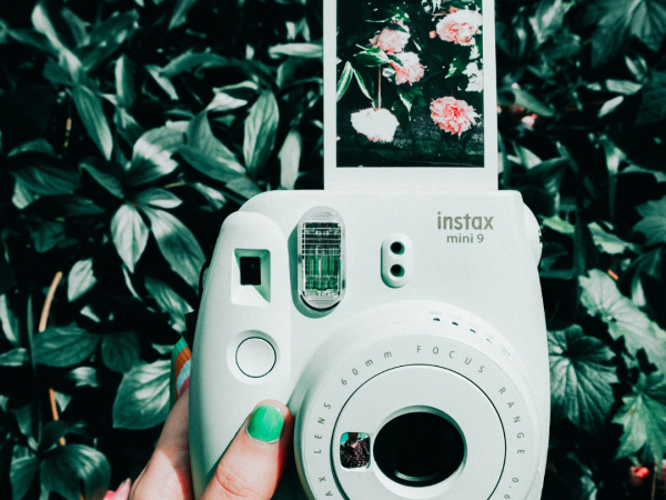 A hand with green nail polish is holding a white Instax camera, displaying a photo of flowers, against a background of lush green leaves and flowers.