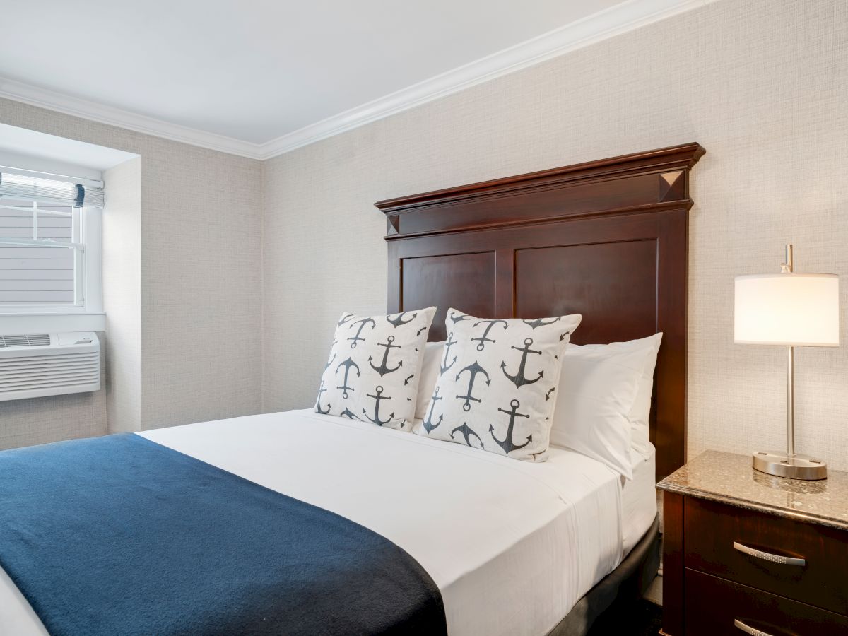 A neatly made bed with anchor-patterned pillows, a blue throw blanket, a wooden headboard, a side table with a lamp, and a window air conditioner.