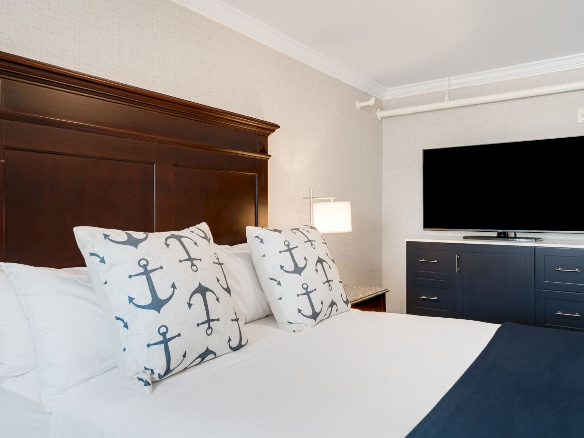 The image shows a well-made bed with nautical-themed pillows, a wooden headboard, a modern dresser with a flat-screen TV, and a table lamp on the side.