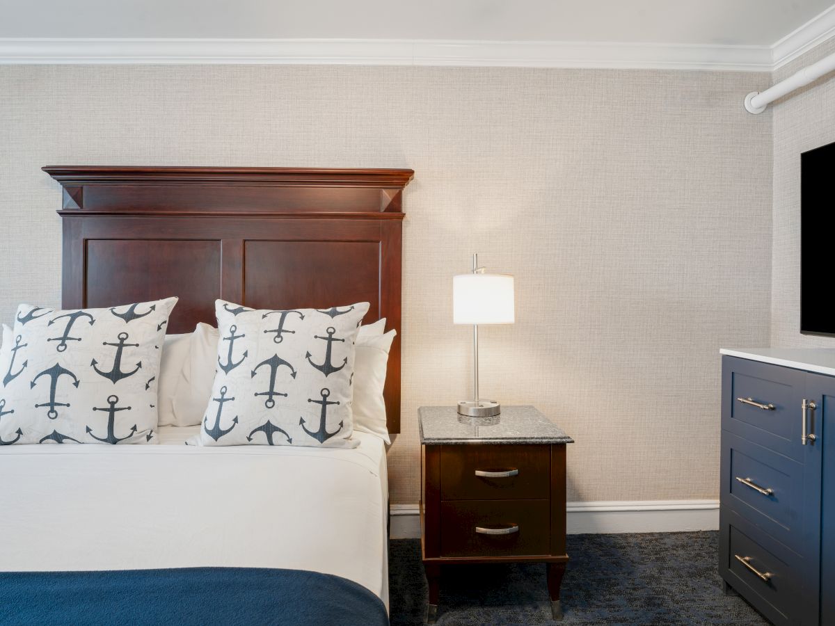 A bedroom with an anchor-themed pillow, a wooden headboard, a bedside table with a lamp, and a dresser with a TV on top.