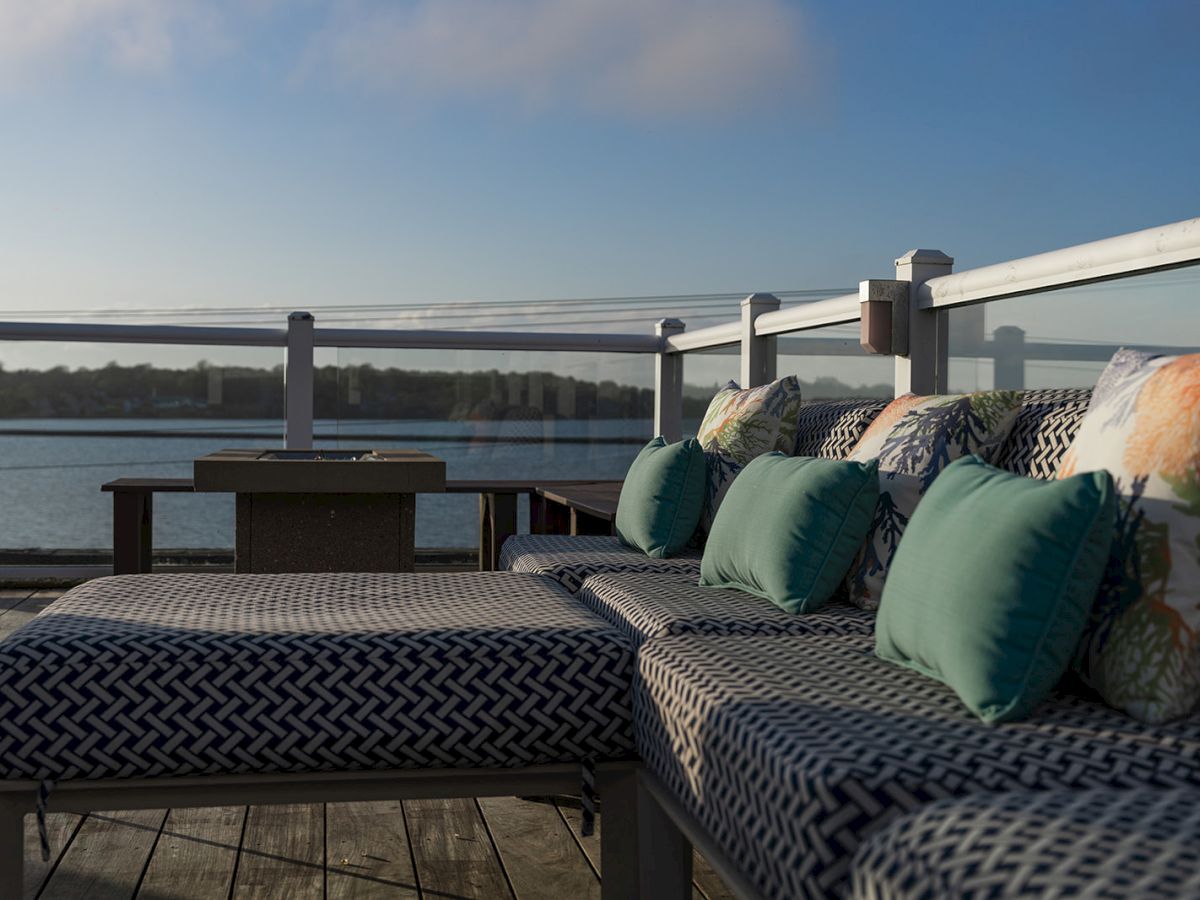 Outdoor lounge area with cushioned seats and pillows overlooking a body of water, surrounded by a glass railing, under a clear sky.