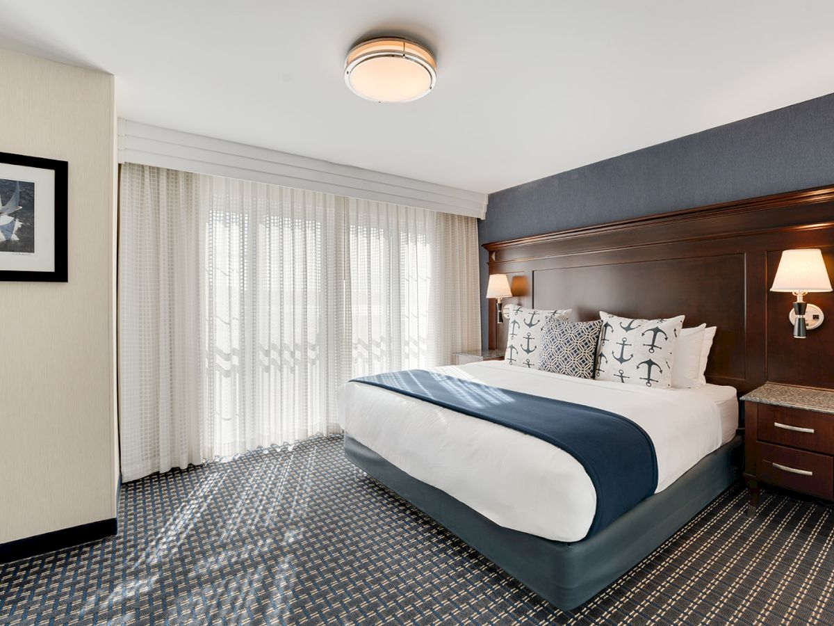 A modern hotel room features a bed with nautical-themed pillows, two bedside tables with lamps, and a large window with sheer curtains.