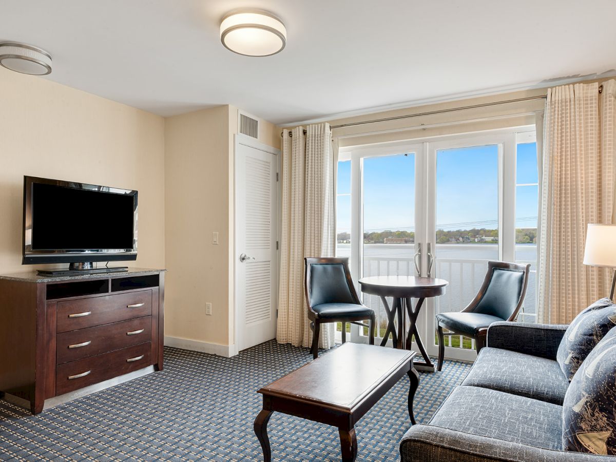 A cozy living room features a couch, a coffee table, a TV on a dresser, two chairs with a small table by a window, and balcony doors with a scenic view.