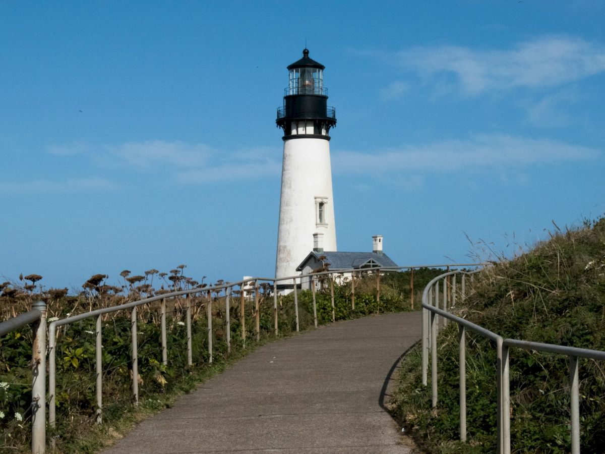 A paved path with railings leads to a tall white lighthouse against a blue sky, surrounded by green vegetation and wildflowers.