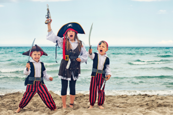 Three children dressed as pirates, holding swords and a toy gun, stand on a sandy beach with the ocean in the background, looking excited.