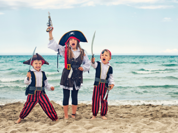 Three children dressed as pirates, holding swords and a toy gun, stand on a sandy beach with the ocean in the background, looking excited.