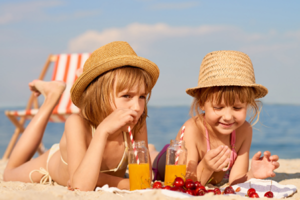 Two children wearing straw hats lie on the beach with drinks and cherries, enjoying a sunny day on a laid-out towel with a sun chair behind them.