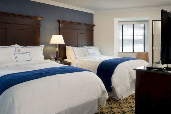 A hotel room with two double beds, white bedding, blue runners, a nightstand with a lamp, a TV, and a window with closed blinds.