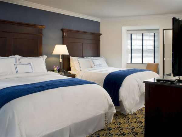 A hotel room with two double beds, white bedding, blue runners, a nightstand with a lamp, a TV, and a window with closed blinds.
