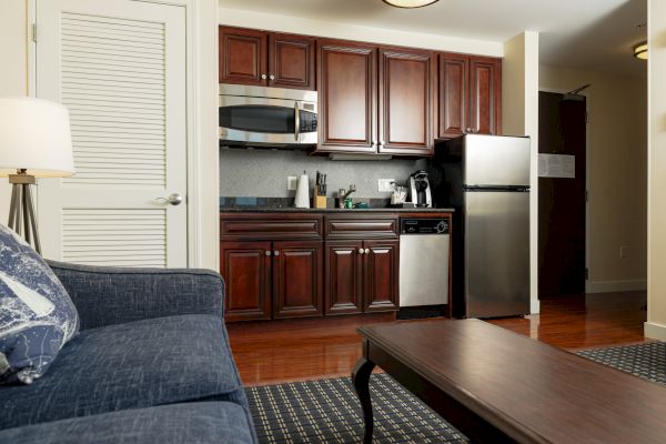 A cozy apartment kitchen features dark wood cabinets, stainless steel appliances, a blue sofa, and a wooden coffee table in a well-lit room.