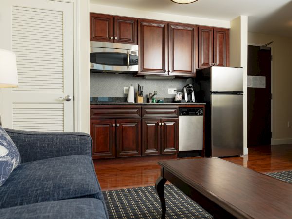 A cozy apartment kitchen features dark wood cabinets, stainless steel appliances, a blue sofa, and a wooden coffee table in a well-lit room.