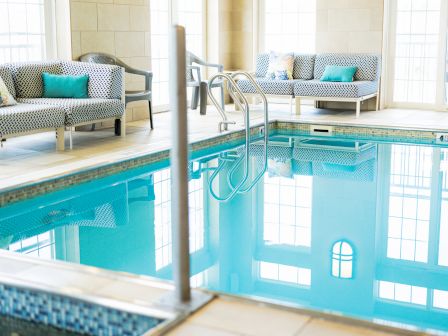 An indoor pool area with clear water, surrounded by cushioned seating and windows allowing natural light to filter in, creating a serene atmosphere.