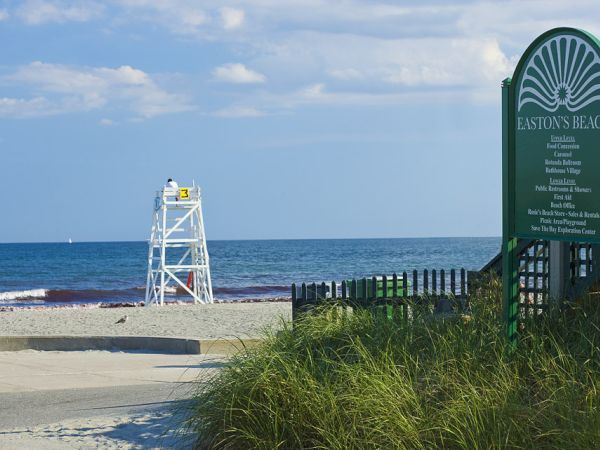A lifeguard tower overlooks the beach with a sign for Easton's Beach to the right, listing amenities and rules. Grass and a path are in the foreground.