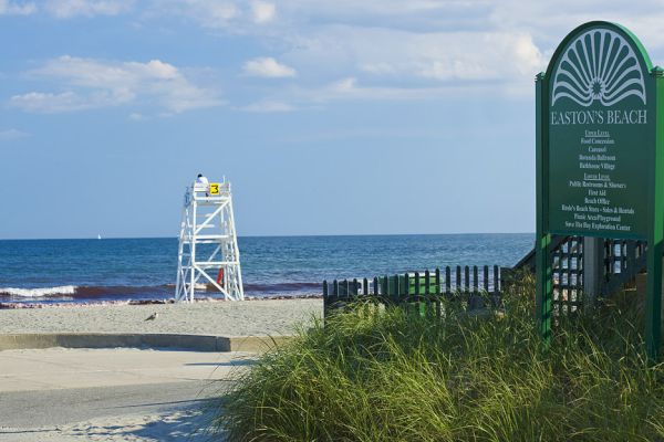 A lifeguard tower overlooks the beach with a sign for Easton's Beach to the right, listing amenities and rules. Grass and a path are in the foreground.