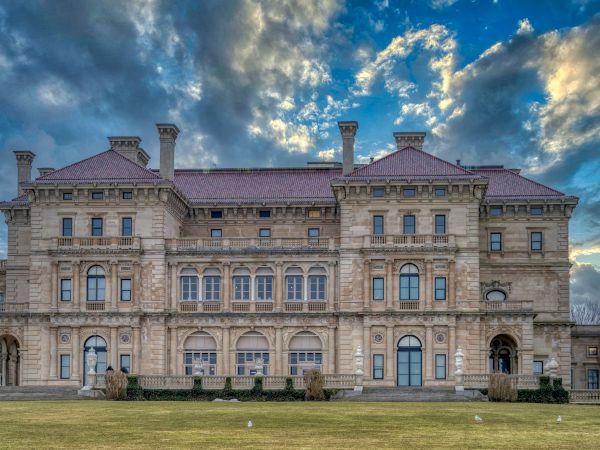The image shows a large, ornate building with intricate architecture against a partly cloudy sky, likely a mansion or historical estate.