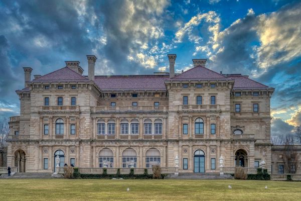 The image shows a large, ornate building with intricate architecture against a partly cloudy sky, likely a mansion or historical estate.