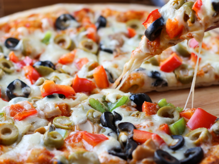 The image shows a vegetable pizza topped with ingredients like black olives, green olives, bell peppers, and melted cheese being served.