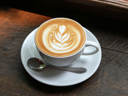 This image shows a cup of coffee with latte art on top, placed on a white saucer with a spoon, set on a wooden surface.