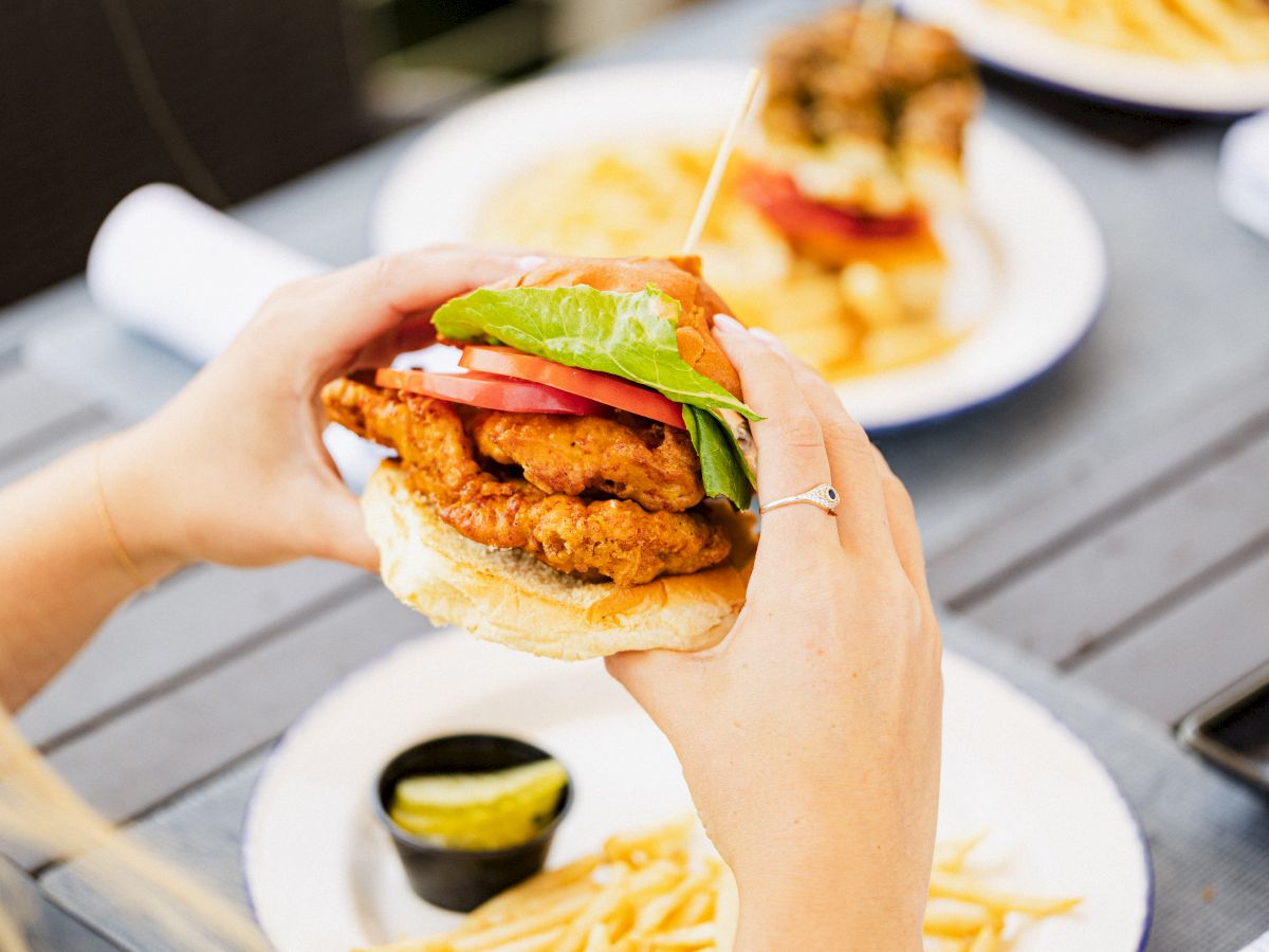 A person is holding a fried chicken sandwich with lettuce and tomato. Plates of fries and pickles are on the table in the background.