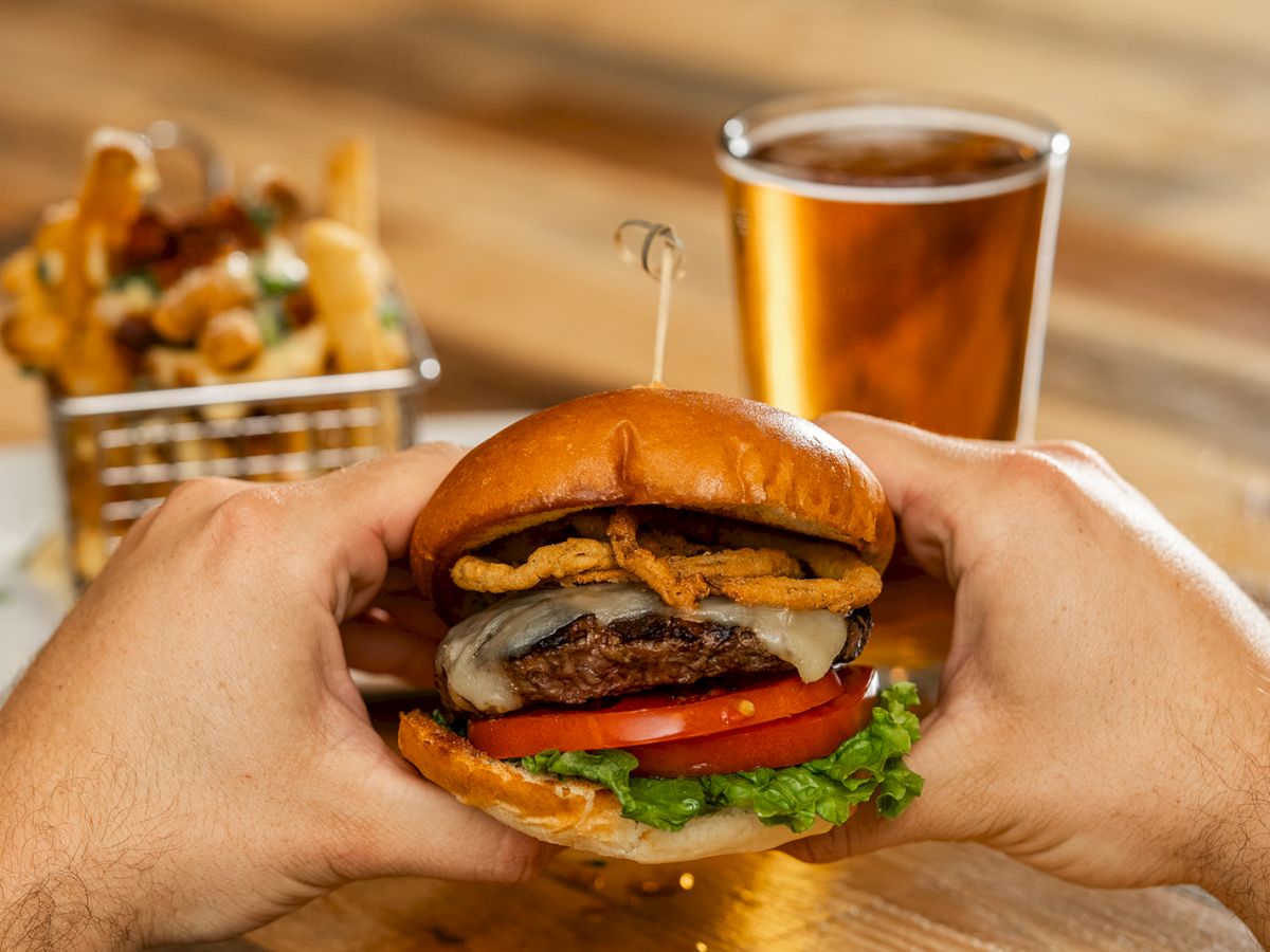 A person holds a burger with lettuce, tomato, cheese, and onions. In the background, there are fries in a basket and a glass of beer.