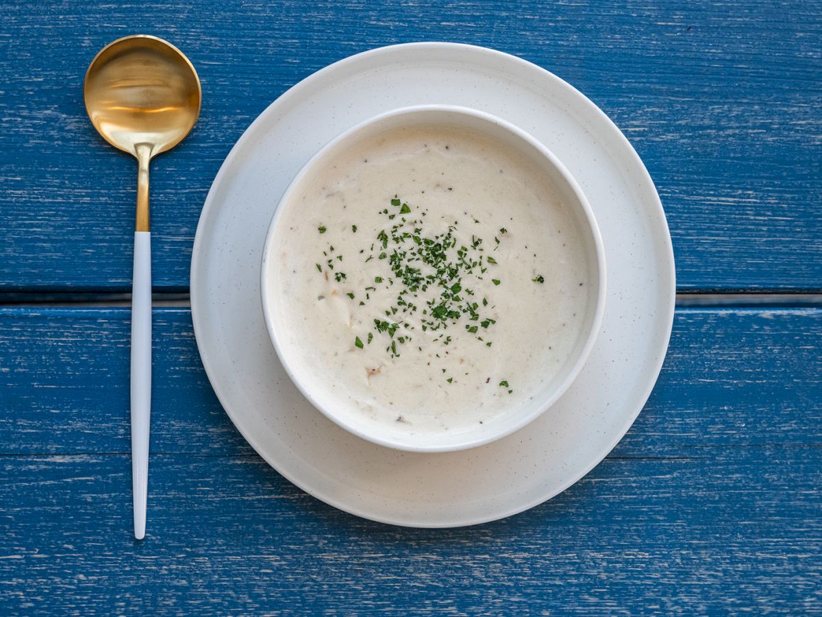 A bowl of creamy soup garnished with herbs on a white plate next to a gold and white spoon, all placed on a blue wooden surface.