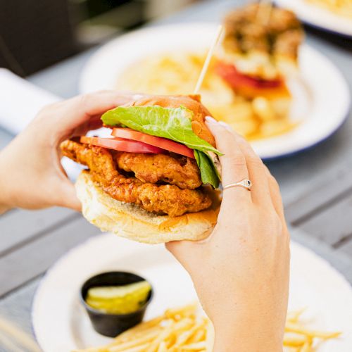 A person is holding a fried chicken sandwich with lettuce and tomato, with a plate of fries and a pickle slice in the background.