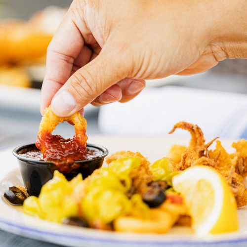 A hand dips a fried calamari ring into sauce on a plate with more calamari, vegetables, and a lemon wedge.