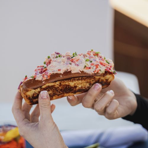 A person is holding a lobster roll over a table with fries and a drink in the background.